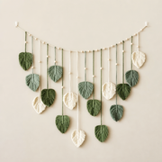 A bohemian-style leaf garland featuring textured macrame leaves in shades of forest green and cream, hanging from a string with small wooden beads against a warm beige wall.