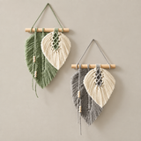 Two bohemian leaf wall hangings displayed on a neutral beige wall. Each hanging features a light wood dowel, knotted cotton cords, and tassels.