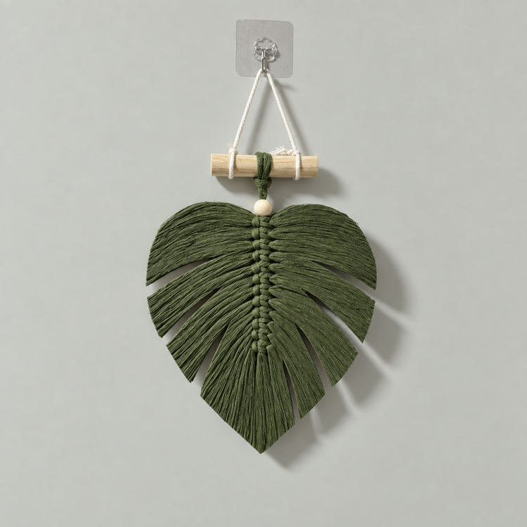 An olive green macrame wall hanging shaped like a monstera leaf, suspended from a light wood dowel with a clear wall hook.