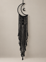A black crescent moon dreamcatcher with intricate woven webbing and long hanging tassels, suspended against a plain light beige wall.