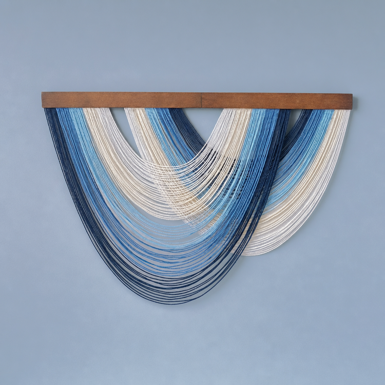 A modern wall art piece featuring multiple curved strands of thread in shades of blue, navy, cream, and white, suspended from a horizontal wooden bar against a solid light blue-gray wall.
