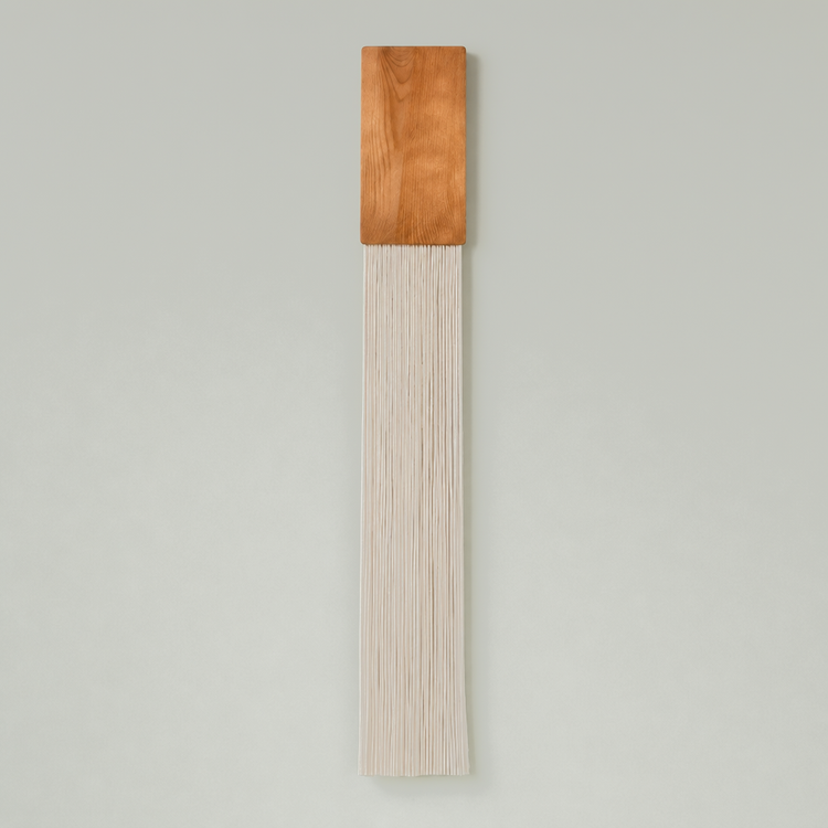 A minimalist wall hanging featuring a rectangular warm brown wood block at the top and a long, narrow fringe of off-white cords below, set against a solid light gray background.
