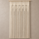 Cream macrame wall hanging with intricate woven patterns and long fringe, suspended from a light wood rod against a neutral beige wall.