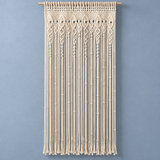 A cream macrame wall hanging with intricate woven patterns and long fringe, suspended from a wooden rod against a solid blue-gray wall.
