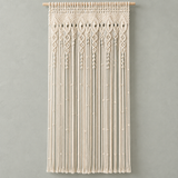 Cream macrame wall hanging with intricate woven patterns and long fringe, suspended from a light wood rod against a textured gray wall.
