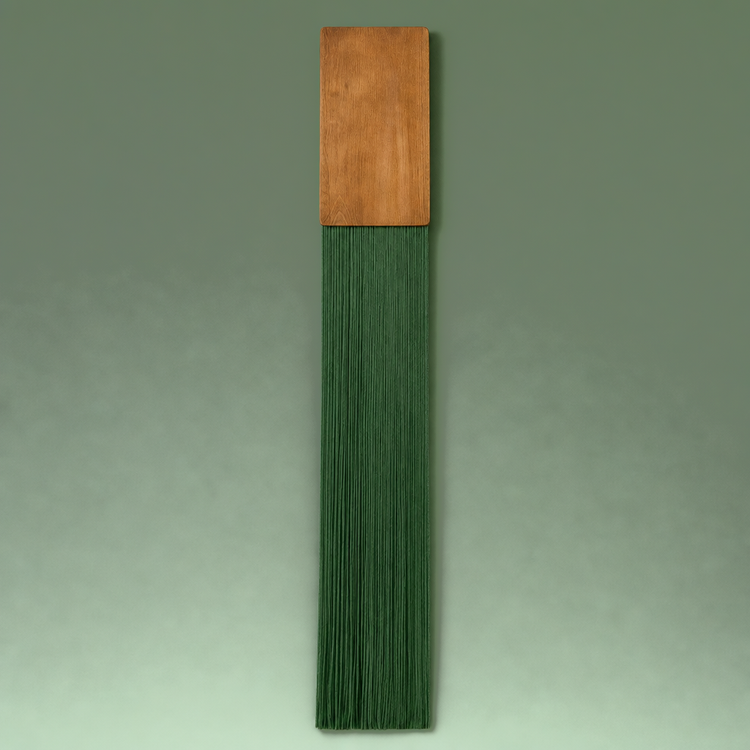 A vertical decorative wall hanging or brush-like decor piece with a rectangular warm brown wooden top and a long, narrow body made of tightly packed dark green strands, shown against a plain green gradient background.