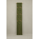 An olive green macrame wall hanging with geometric diamond patterns and a long fringe, suspended from a light natural wood dowel against a neutral gray studio background.