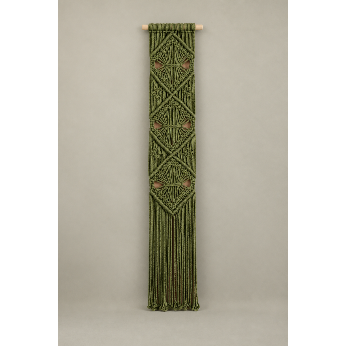 An olive green macrame wall hanging with geometric diamond patterns and a long fringe, suspended from a light natural wood dowel against a neutral gray studio background.