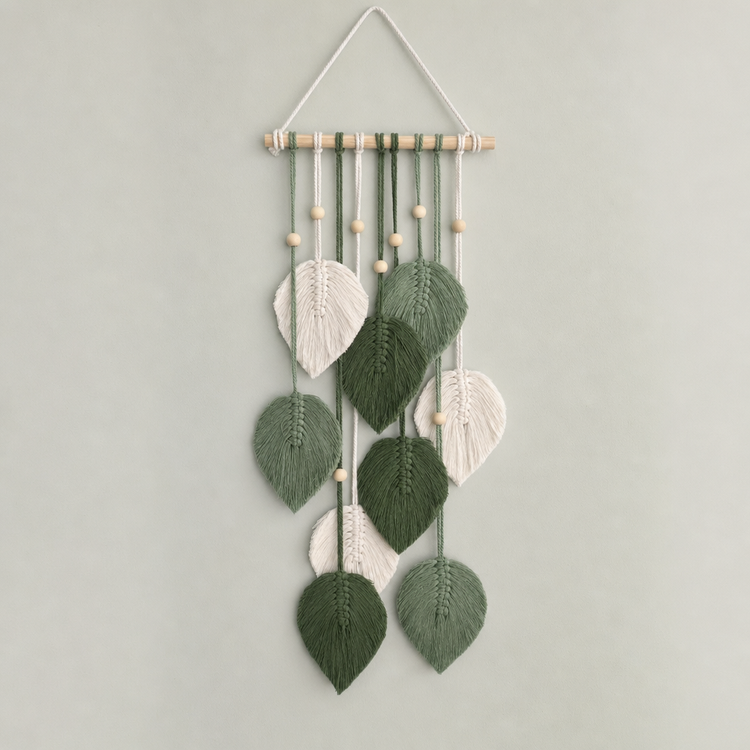 A hanging wall decor piece made of multiple embroidered leaf shapes in shades of green and off-white, suspended from a light wood dowel with small wooden beads, against a plain light gray wall.