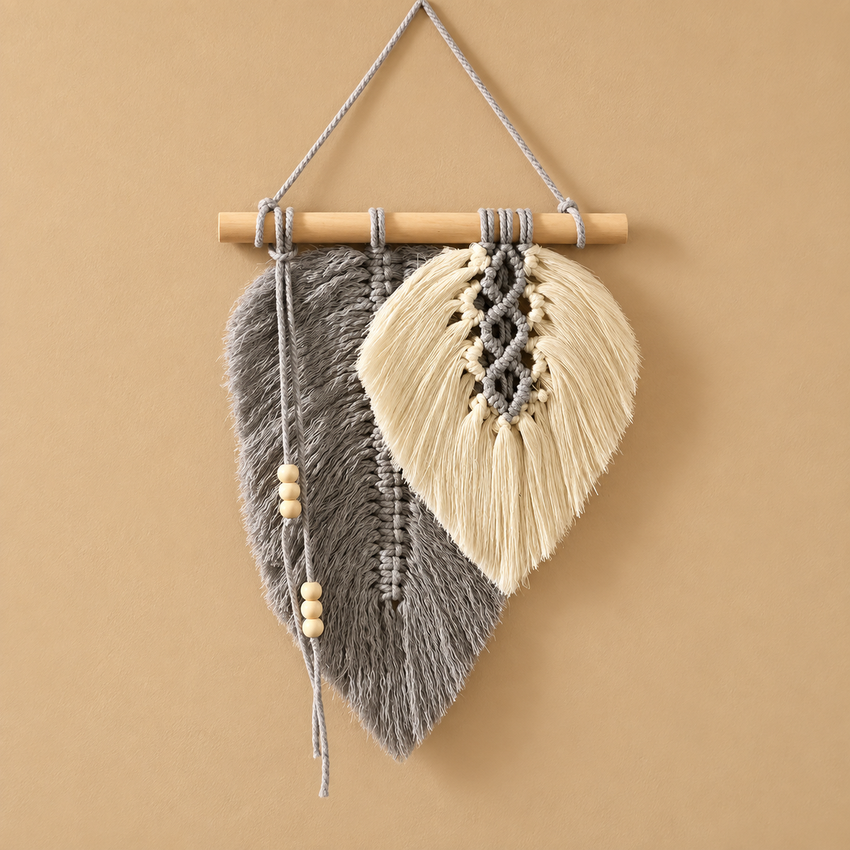 A bohemian-style woven wall hanging featuring two leaf-shaped panels, one in gray and one in cream, suspended from a light wood dowel with a gray hanging cord against a solid tan background.