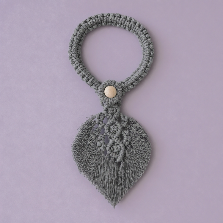A sage green macrame bracelet with a textured leaf-shaped tassel and a small gold button accent, displayed on a solid lavender background.
