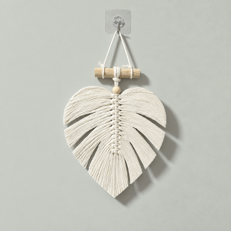 A white woven macrame wall hanging shaped like a monstera leaf, suspended from a light wood dowel with a string on a warm off-white background.