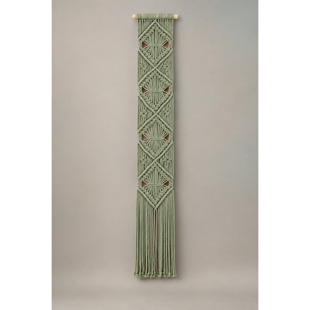 A vertical macrame wall hanging in sage green with geometric diamond patterns and small brown accents, suspended from a light wood dowel against a neutral gray background.