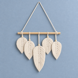 A bohemian-style macrame leaf wall hanging with five woven cream cotton leaves suspended from a light natural wood dowel, hung by a matching cream rope against a solid dusty blue background.