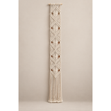 A vertical cream macrame wall hanging with geometric diamond patterns and long fringe, suspended from a light wooden rod against a plain beige studio background.