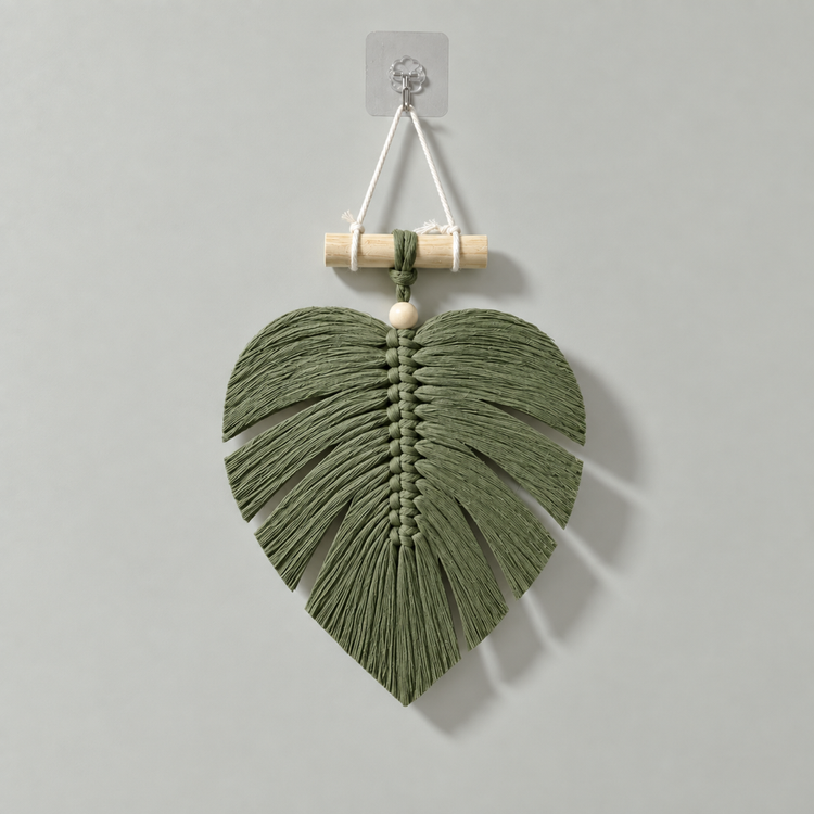 An olive green macrame wall hanging shaped like a monstera leaf, suspended from a wooden dowel with a clear wall hook on a light gray background.