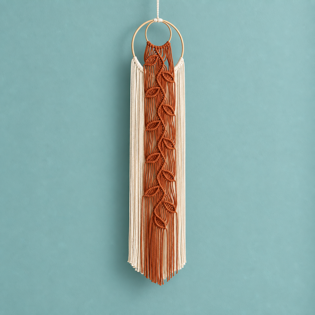 A bohemian-style macrame wall hanging suspended from a light wood ring, featuring a central rust-brown woven leaf pattern over cream-colored fringe, set against a solid teal background.