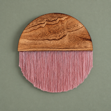 A circular wall hanging with a natural wood top half and pink fringe hanging from the bottom, shown against a muted sage green background.