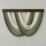 A modern wall hanging sculpture featuring two interlocking arches of layered cord in dark and light olive green and cream, suspended from a dark brown wooden bar against a light gray wall.