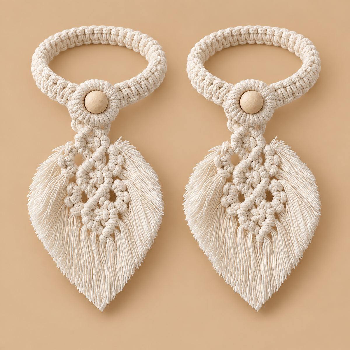 Two matching cream macrame hair ties with leaf-shaped tassels and wooden button centers, displayed on a solid beige background.