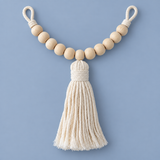 A decorative hanging ornament featuring natural wood beads strung on a cream rope cord with a large cream tassel, shown against a solid periwinkle blue background.