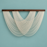 A modern wall hanging sculpture featuring multiple cream-colored cords suspended from a dark brown wooden bar, set against a solid light blue background.