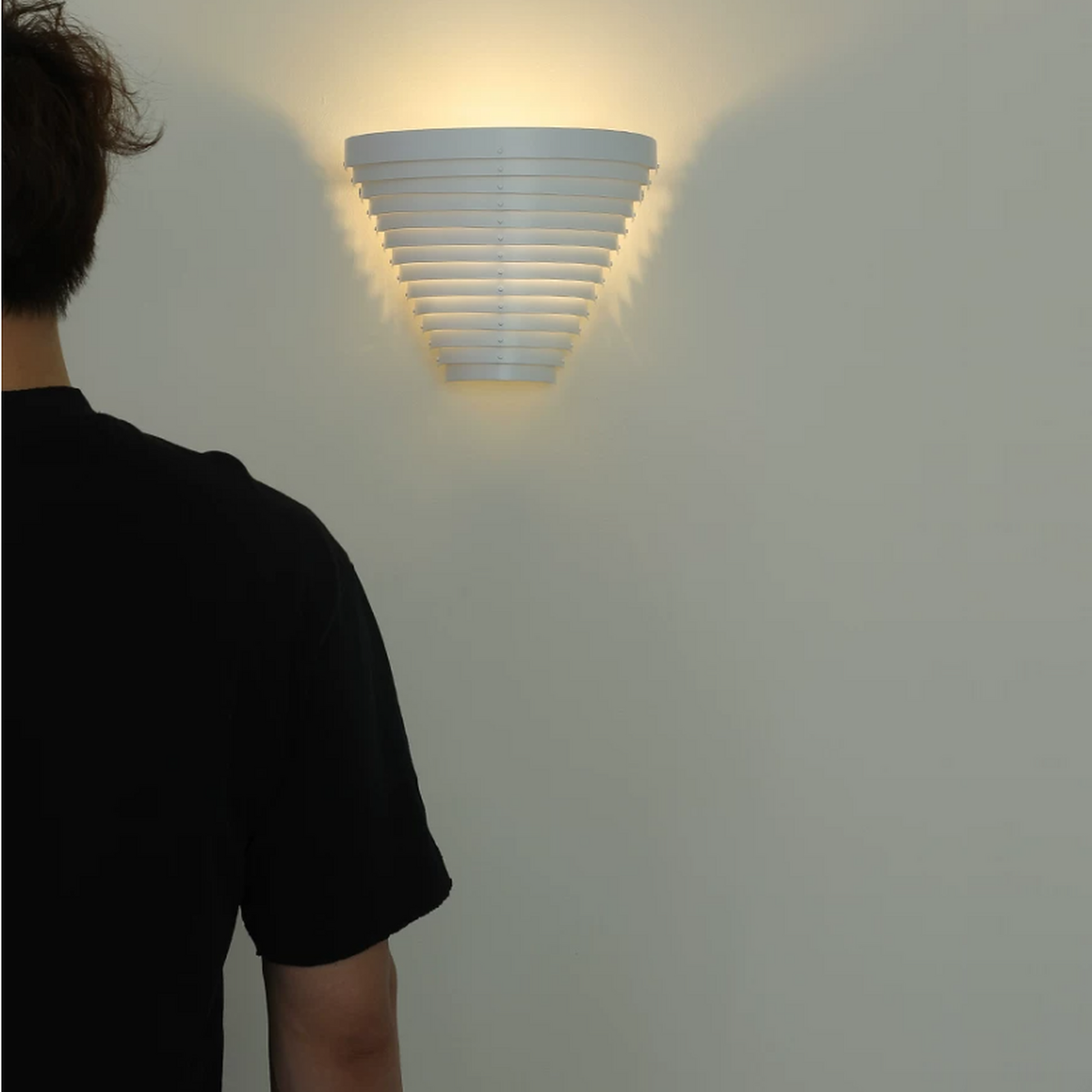 A modern white tiered wall sconce light fixture mounted on a light gray wall, casting a warm yellow glow. A person in a black t-shirt is seen from behind on the left side of the frame.