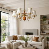 A brass six-light chandelier with white drum shades hangs in a bright, modern living room with a cream sectional sofa, fireplace, and shelving.